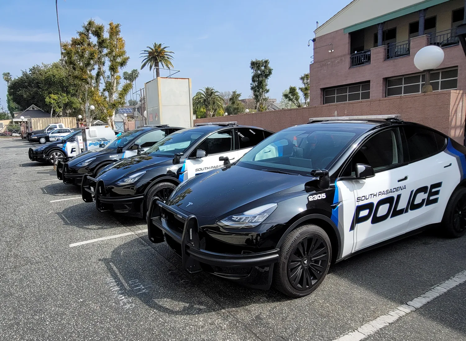 A line of five police cars parked in a lot, with three black vehicles and two white vehicles featuring police markings. In the background are palm trees and a building.