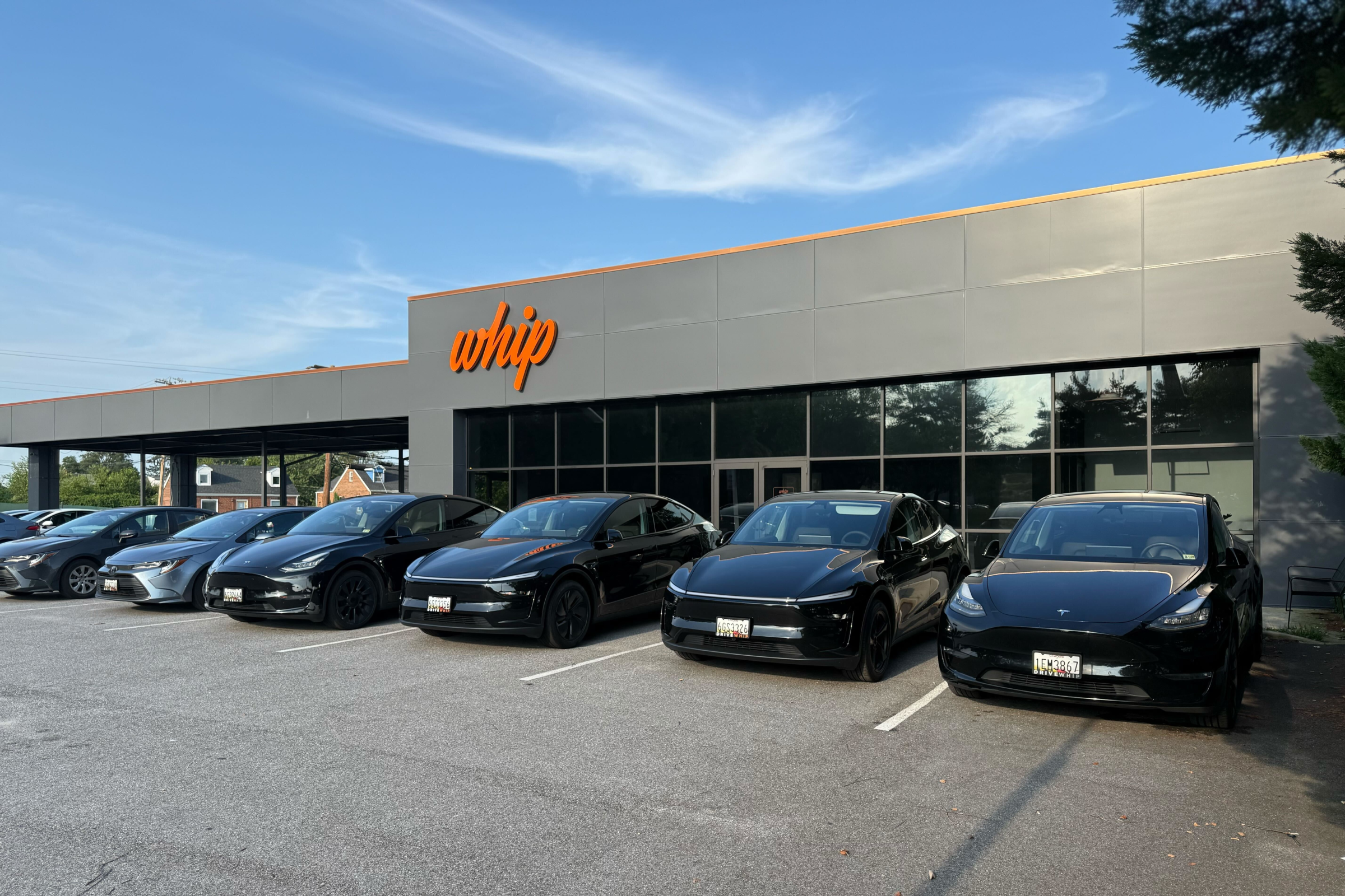 A row of six black Tesla cars parked in front of a modern building with a prominent orange "whip" sign visible on the facade.