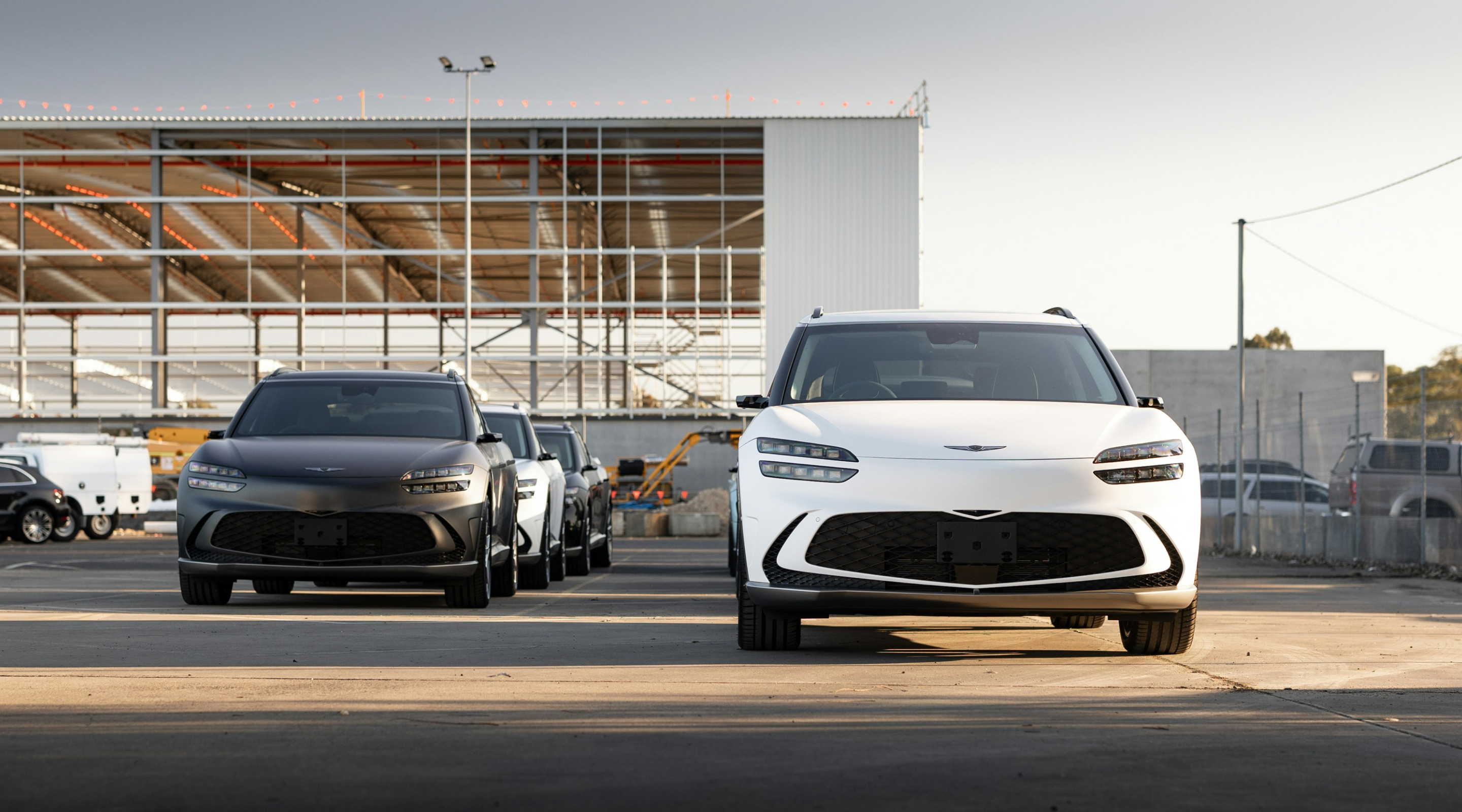 Two Genesis SUVs, one black and one white, parked in front of an industrial building with metal framing. The setting is an outdoor area with construction elements visible in the background.