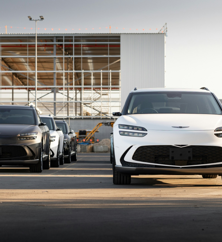 A lineup of modern vehicles, including a white SUV in the foreground and two dark-colored vehicles behind it, parked on a concrete surface with a construction site in the background.