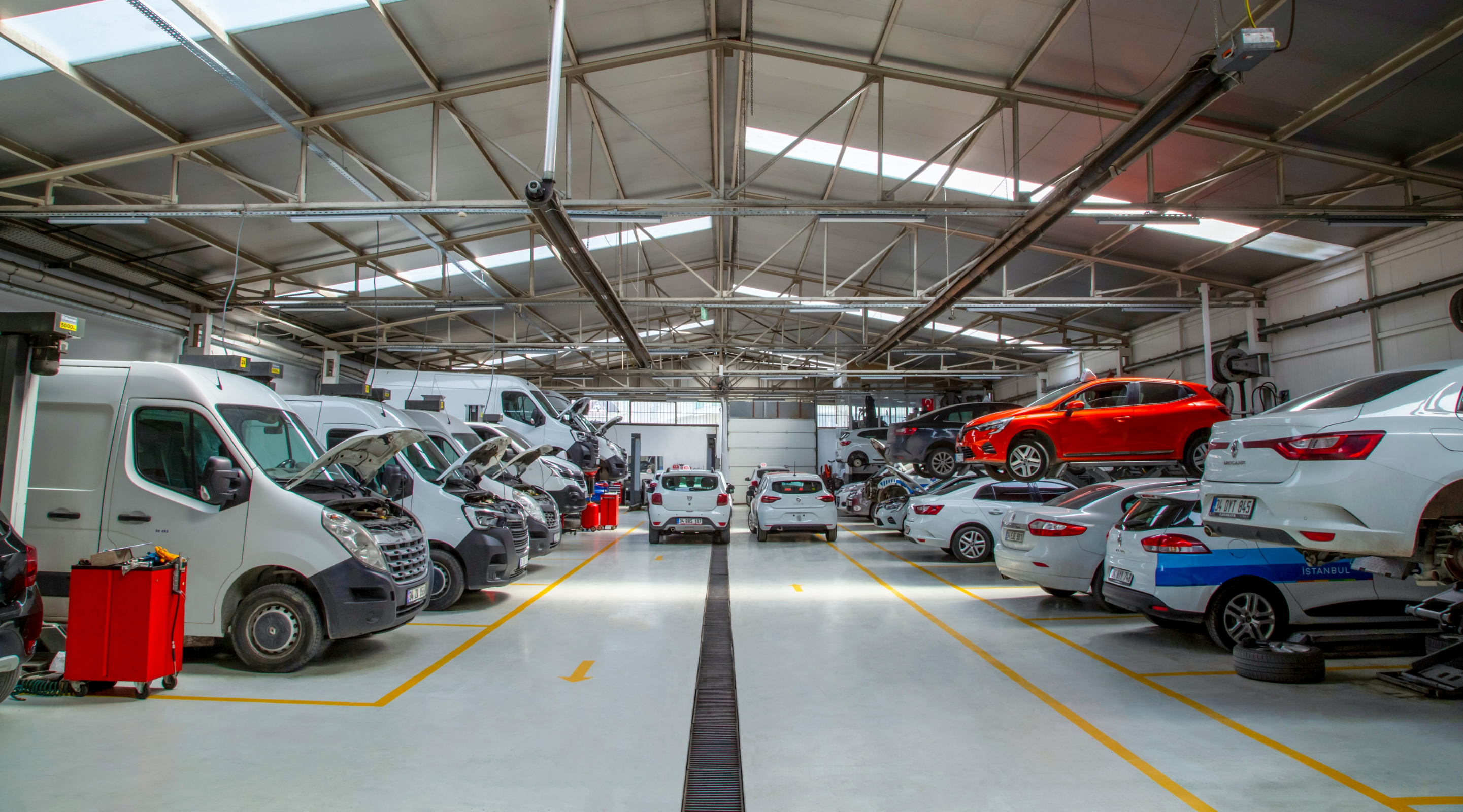 A spacious automotive workshop with various vehicles, including cars and vans, undergoing maintenance. Some cars are elevated on lifts, while others are parked along marked lanes, with bright overhead lights illuminating the interior.