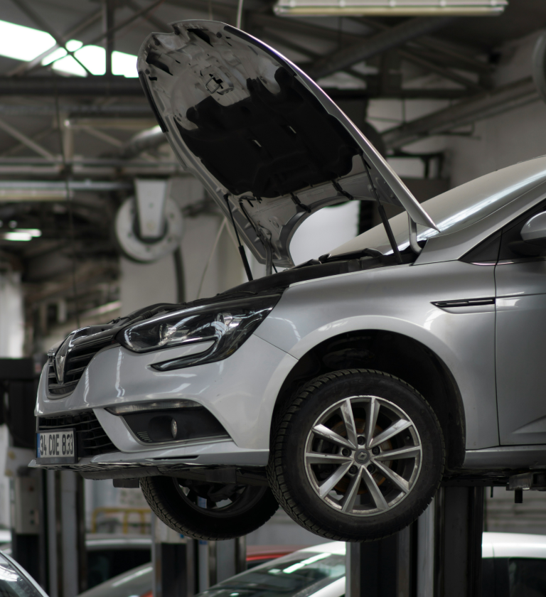 A silver car with its hood raised is positioned on a hydraulic lift in an automotive workshop. The background shows various tools and another vehicle in a well-lit garage.
