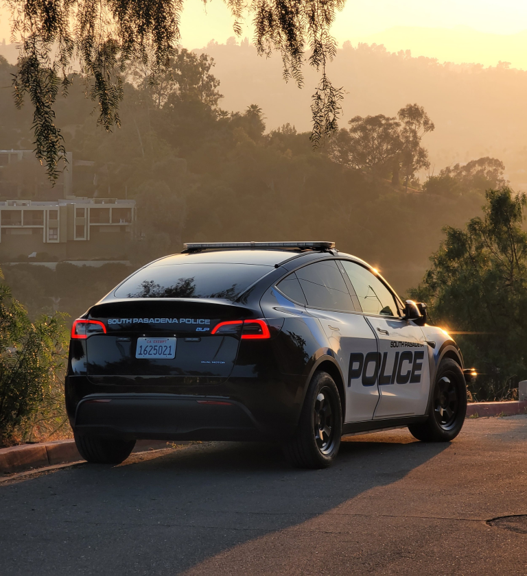 A South Pasadena police vehicle parked on a roadway with a scenic view in the background during sunset. The car features a black and white design with the word 'POLICE' prominently displayed.