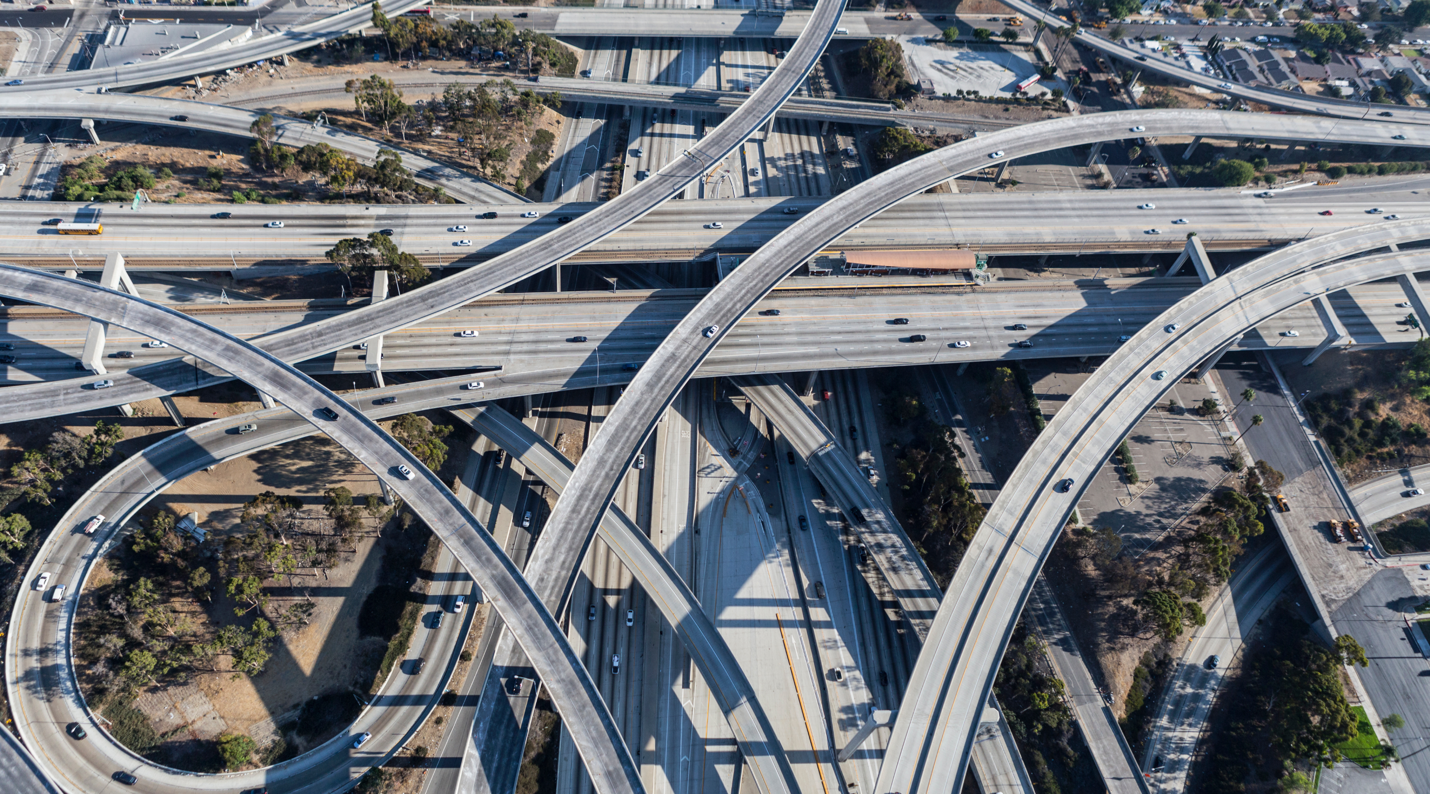 An aerial view of a complex highway interchange with multiple lanes and overpasses, featuring cars traveling in various directions amid green spaces and urban structures in the background.