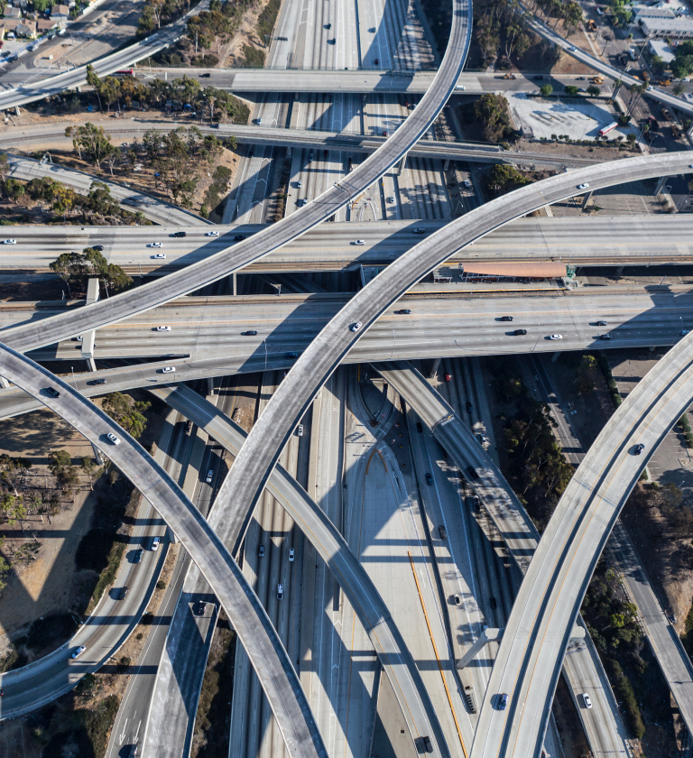 Aerial view of intersecting highways and overpasses, with vehicles traveling on multiple lanes. The scene includes greenery and urban structures in the background.