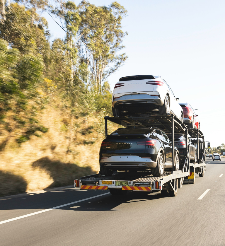A car carrier truck transporting multiple vehicles, including several SUVs in white and grey, driving on a highway surrounded by trees.