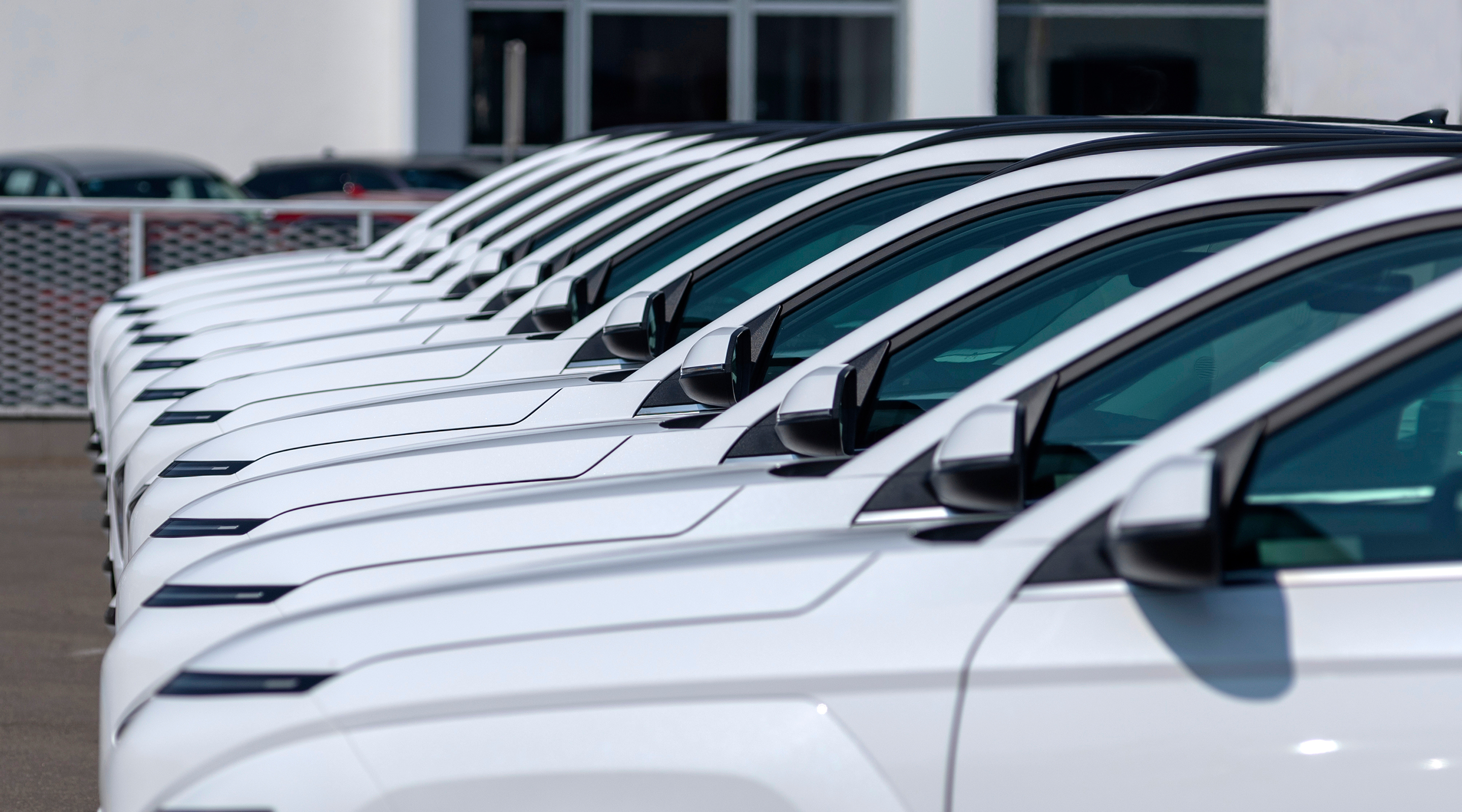 A row of white vehicles parked in a dealership lot, showcasing the side view and mirrors of each car. The background includes more cars parked in the distance.
