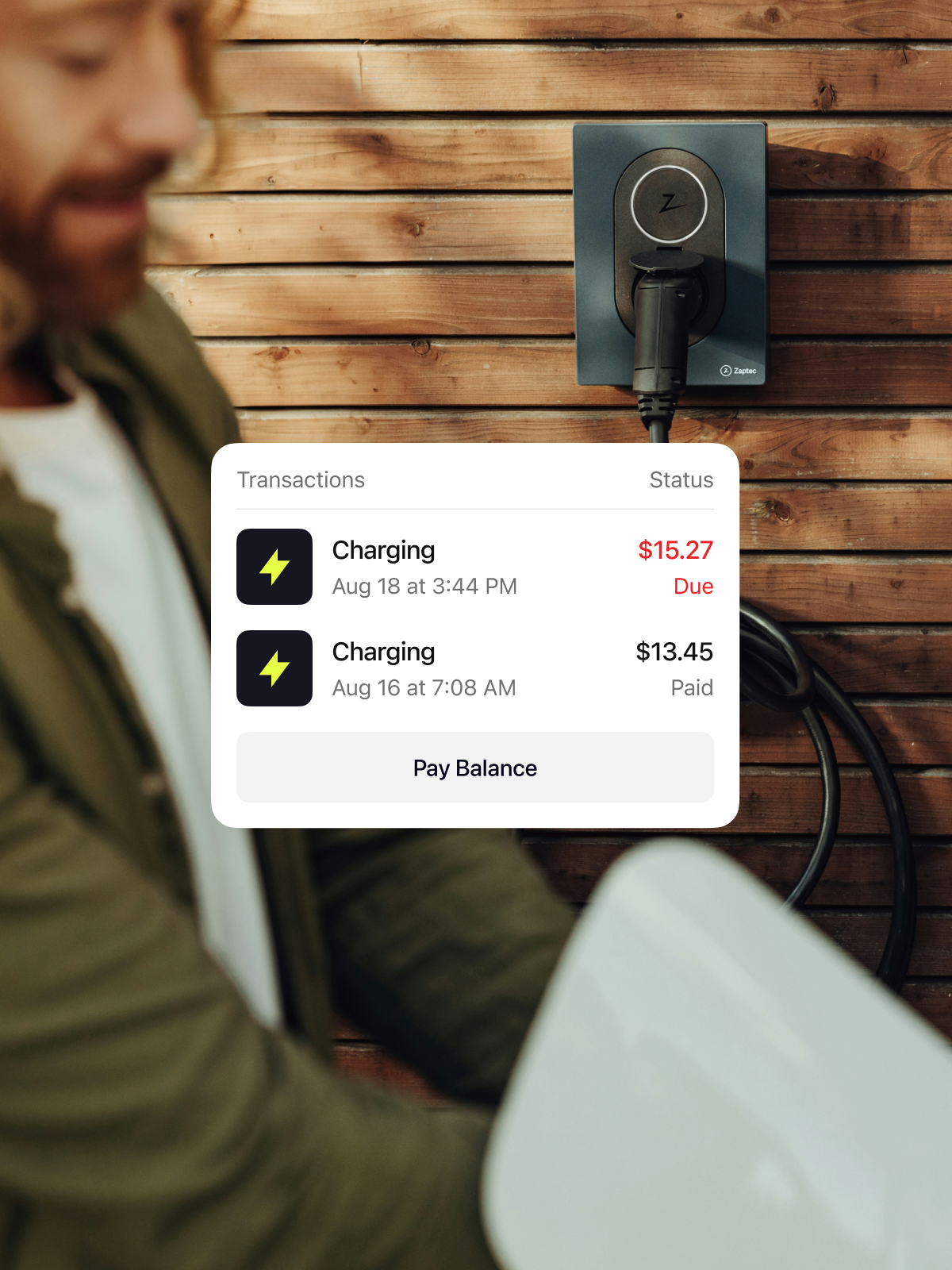 A person smiling while using an electric vehicle charging station mounted on a wooden wall. An app interface displays charging transactions and amounts due.