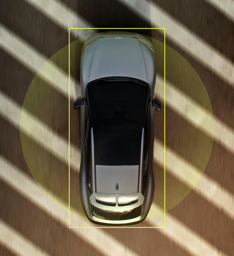 An overhead view of a sleek silver car positioned under striped shadows, highlighting its streamlined design. The car is parked on a smooth surface, surrounded by a circular green backdrop.