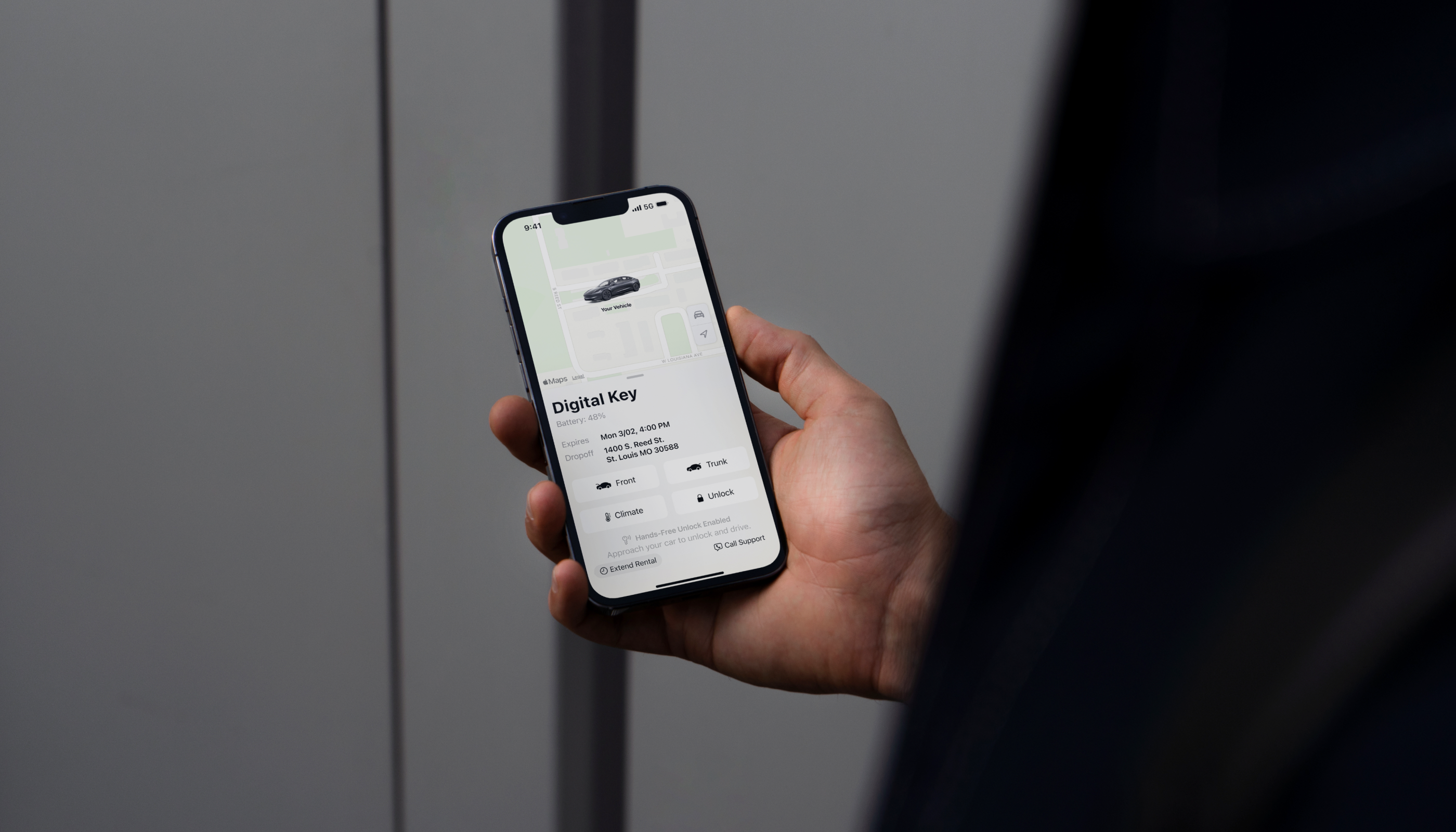 A hand holding a smartphone displaying a digital car key application. The screen shows a vehicle illustration and controls related to the digital key.