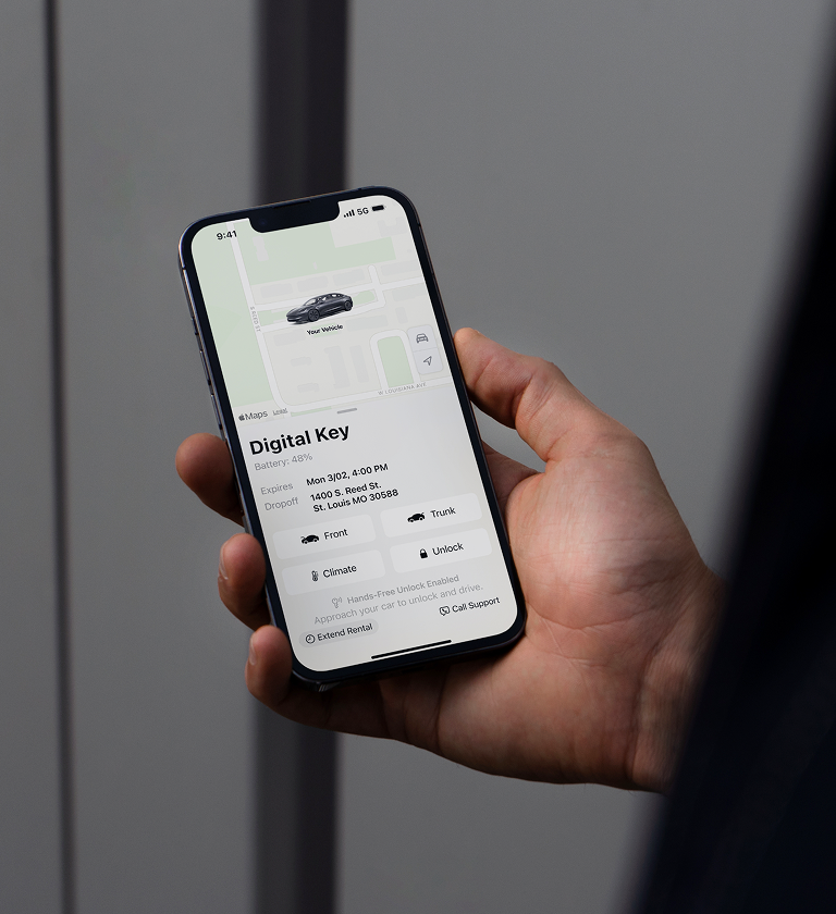 A person holding a smartphone displaying a digital key interface for a car rental. The screen shows vehicle details, including model, pickup time, and location information.