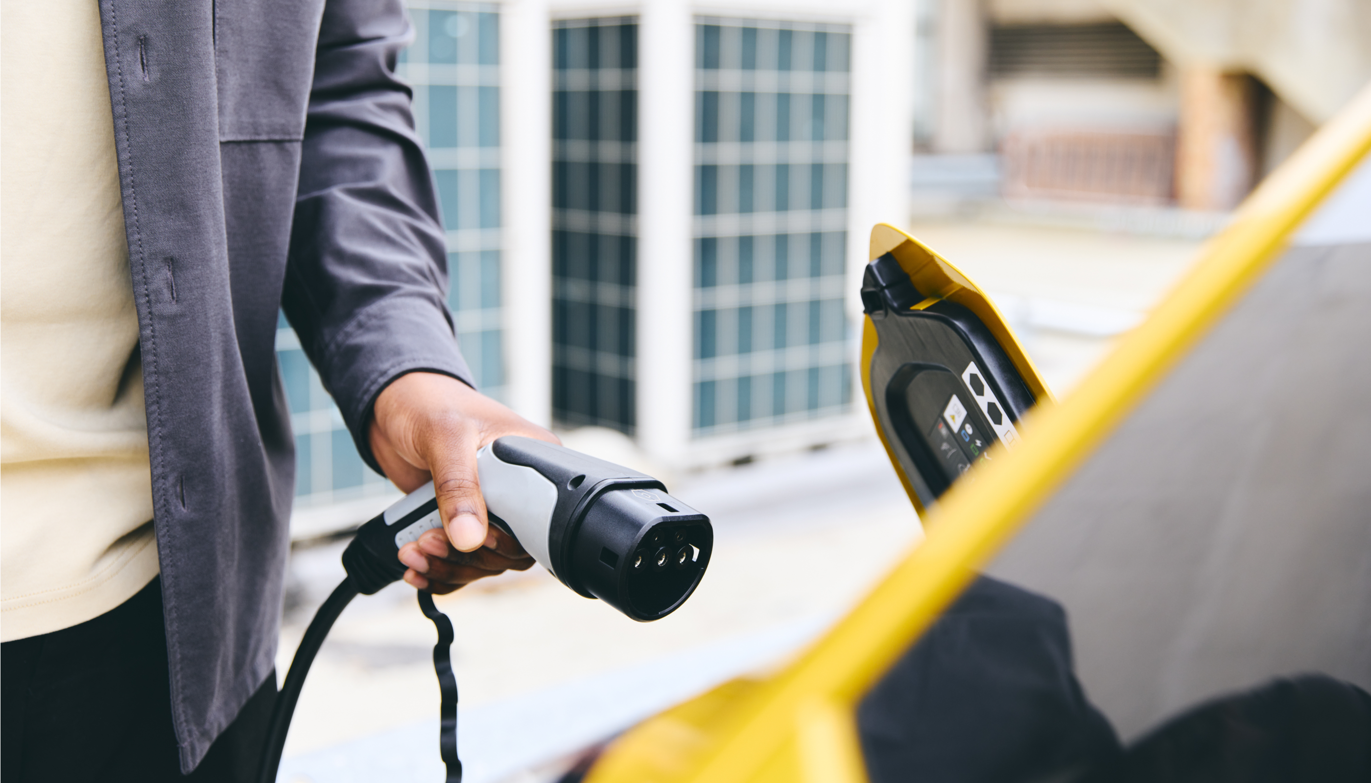 A person holding an electric vehicle charging connector, preparing to plug it into a yellow car. The background features large solar panels and a modern urban setting.