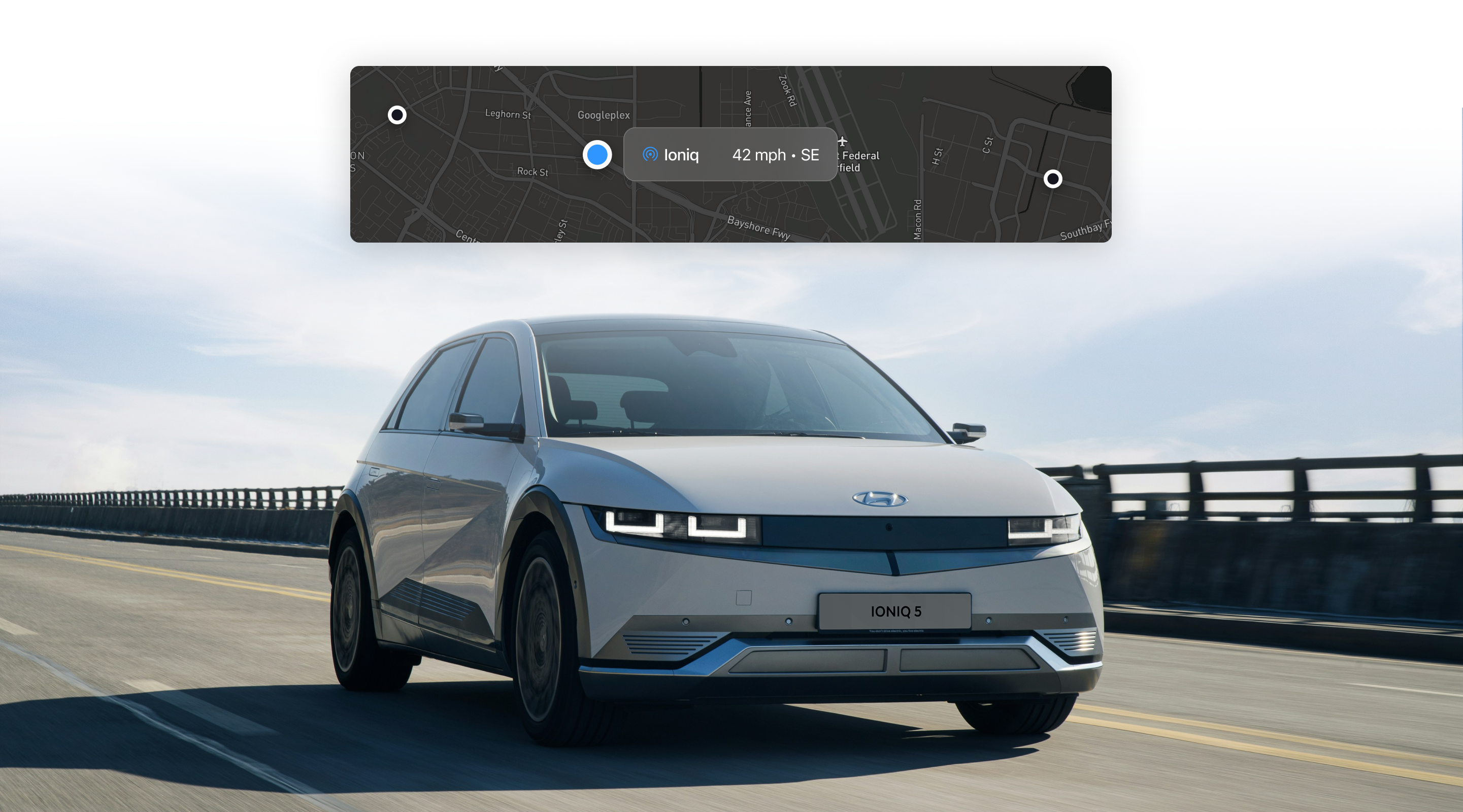 A silver Hyundai Ioniq 5 electric vehicle driving on a road with a scenic backdrop. A digital display shows the vehicle's speed and location on a map.