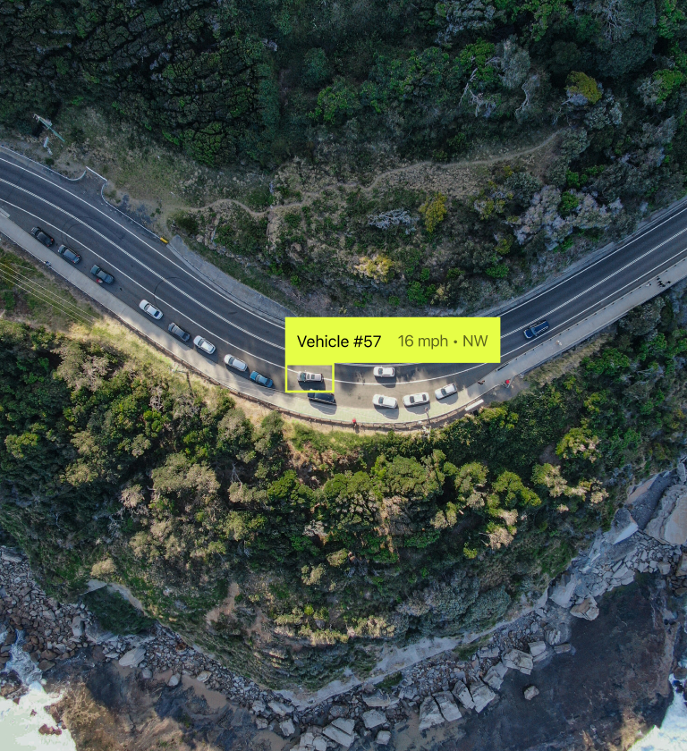 An aerial view of a winding coastal road with several vehicles, including one labeled 'Vehicle #57' moving at 16 mph northwest. The surrounding landscape features green vegetation and rocky shoreline.