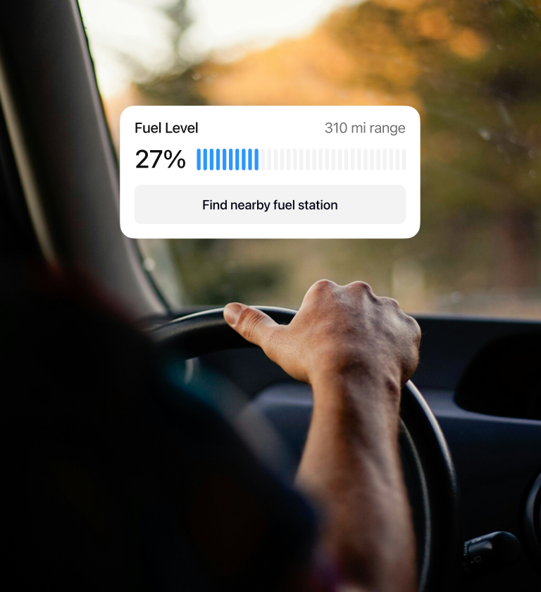 A person's hand on the steering wheel inside a vehicle, with a digital display showing fuel level at 27% and a range of 310 miles, alongside an option to find a nearby fuel station.