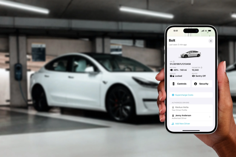 A person holding a smartphone displaying a vehicle management app interface. In the background, a white electric car is parked at a charging station in an underground parking lot.