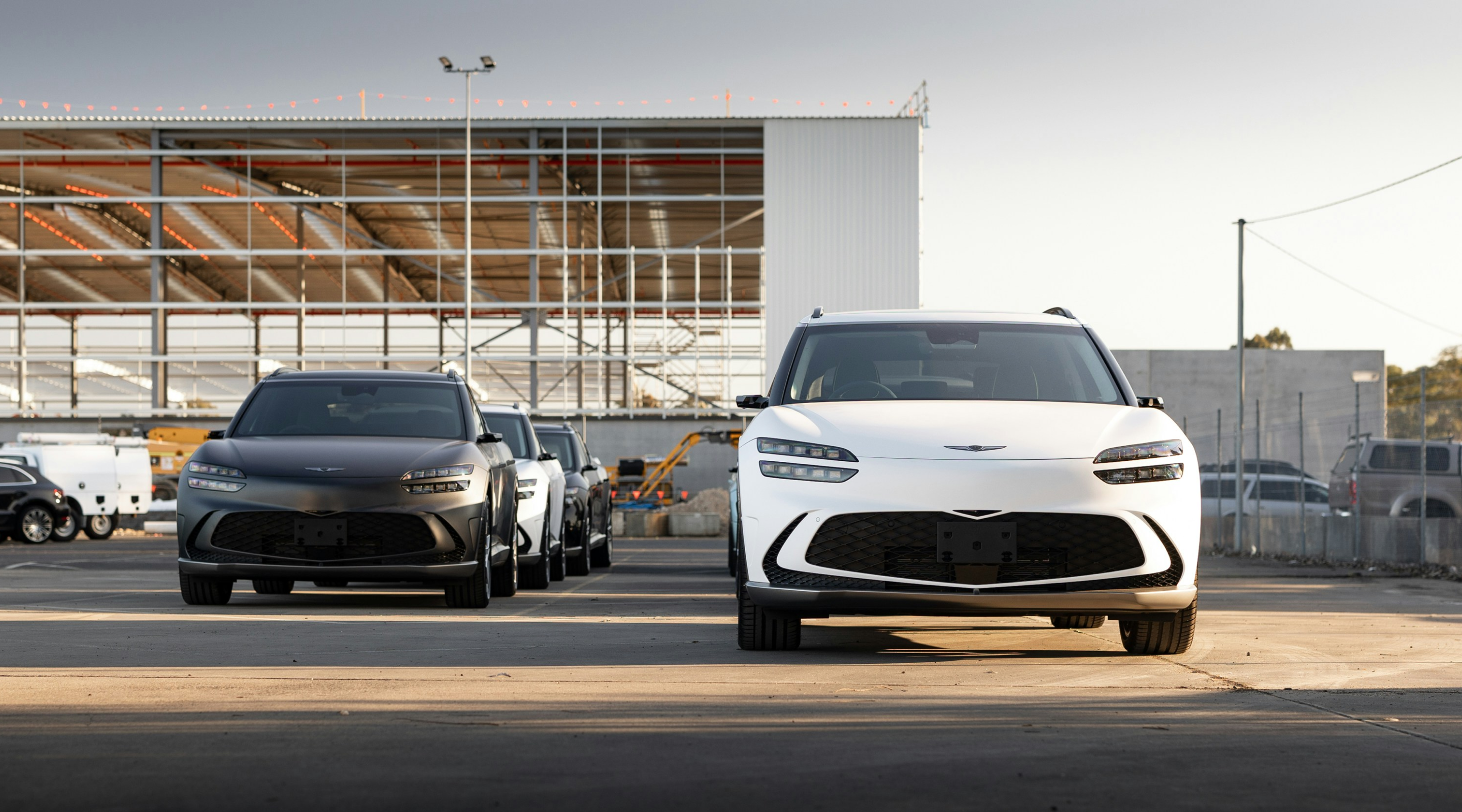 A lineup of three Genesis vehicles, two SUV models in dark gray and white colors, parked in an industrial area, with construction structures in the background under an overcast sky.