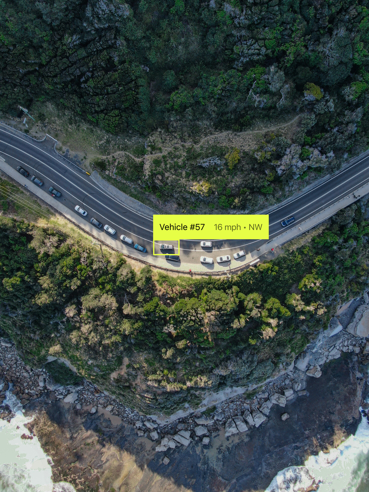 Aerial view of a winding coastal road with several vehicles, including one labeled 'Vehicle #57' moving at 16 mph, near a rocky shoreline and lush greenery.