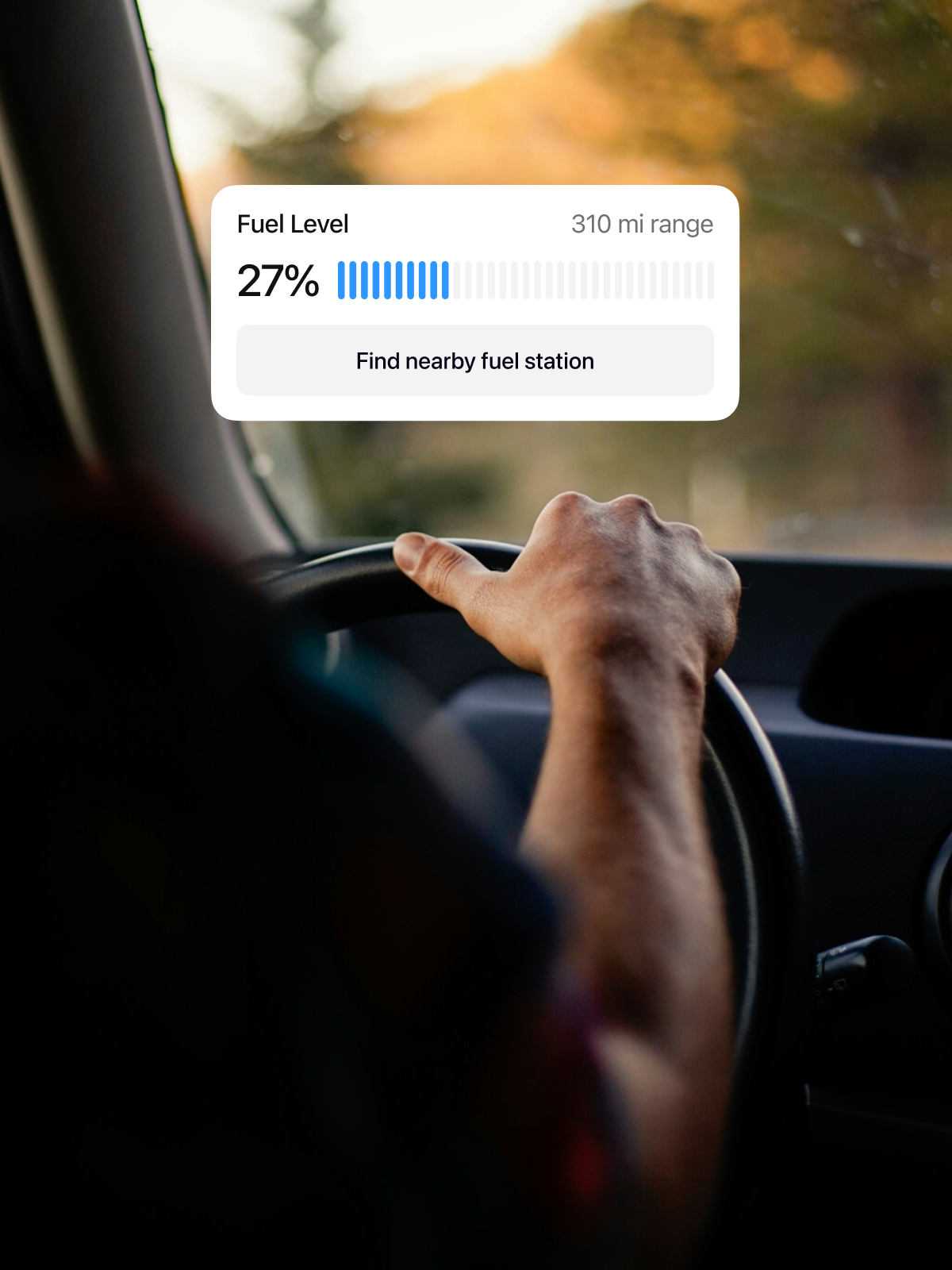 A driver’s hand on the steering wheel inside a car. An interface displays the fuel level at 27%, indicating a 310-mile range and a prompt to find a nearby fuel station.